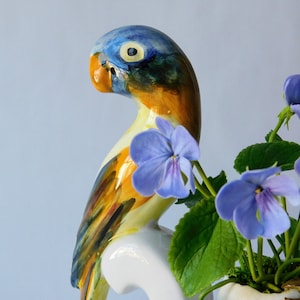 Puede incluir: Una figurita de loro de cerámica pintada en azul, amarillo y naranja, posada sobre una base de cerámica blanca. El loro está rodeado de una pequeña planta en maceta con flores azules.