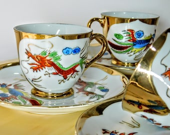 Può includere: Set di quattro tazze da tè e piattini in porcellana bianca con bordi dorati. Ogni tazza presenta un diverso design colorato di ispirazione asiatica, tra cui un drago e una fenice.