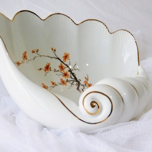 May include: White ceramic serving bowl with a seashell design. The bowl features a scalloped rim with gold trim and a painted floral branch with orange blossoms. The bowl has a spiral handle.