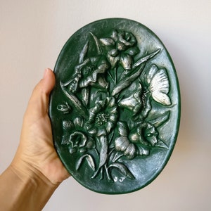 Puede incluir: Un plato de pared ovalado de cerámica verde con un diseño en relieve de flores y hojas plateadas.
