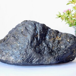 Könnte beinhalten: Ein großer, dunkelgrauer, grob strukturierter Stein mit einem leicht metallischen Schimmer. Der Stein hat eine abgerundete, unregelmäßige Form.