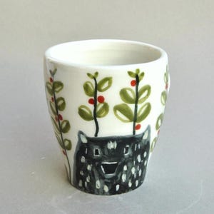 Può includere: Un bicchiere in ceramica bianca con un'illustrazione di orso nero alla base. Foglie verdi e bacche rosse sono dipinte sulla superficie bianca. Il bicchiere è cilindrico con un bordo leggermente svasato.