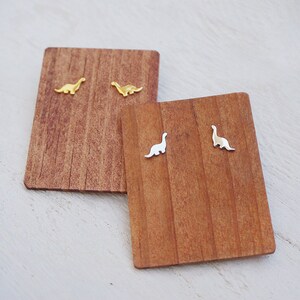 May include: Two pairs of dinosaur stud earrings on wooden display cards. One pair is gold and the other is silver.