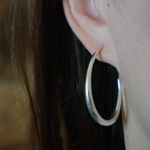 May include: Silver hoop earrings worn in a person's earlobe. The earrings are large and have a smooth, polished finish.