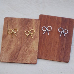May include: Two pairs of bow-shaped earrings displayed on a wooden board. One pair is gold and the other is silver.