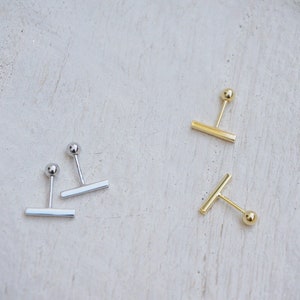 May include: Two pairs of minimalist earrings with a small ball and a horizontal bar. One pair is silver and the other is gold.