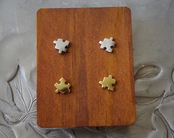May include: Two pairs of puzzle piece earrings, one pair is silver and the other is gold. The earrings are displayed on a wooden surface.