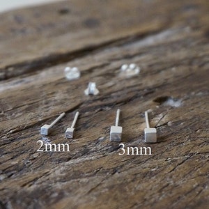 Puede incluir: Pendientes cuadrados de plata, de 2 mm y 3 mm, presentados sobre una superficie de madera rústica. La imagen incluye cierres y detalles de tamaño. Diseño minimalista.