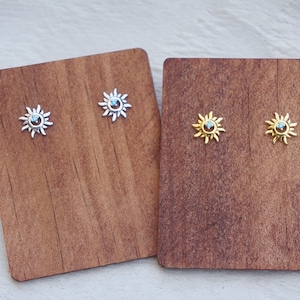 May include: Two pairs of earrings on a wooden display. The earrings are small and feature a sun design with a blue gemstone in the center. One pair is silver and the other is gold.