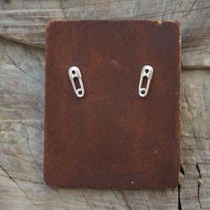 May include: A pair of silver safety pin earrings displayed on a rectangular, brown leather backing. The earrings are small and detailed, with a classic safety pin design. The leather has a textured appearance, and the background is a wooden surface.
