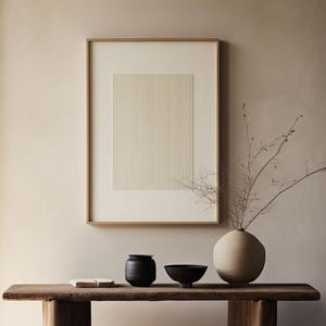 May include: A rectangular wooden console table with two thick legs supports a small white book, a black cylindrical vase, a black bowl, and a large round beige vase containing thin branches. A framed print with vertical lines hangs above.