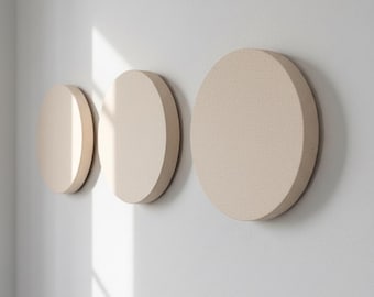 May include: Three beige, circular wall decor pieces of varying sizes are mounted on a white wall. The objects have a textured surface and are arranged in a row, creating a minimalist aesthetic. The lighting casts shadows.