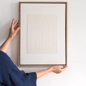 May include: A framed, abstract artwork with a white, textured surface featuring vertical and square patterns. The artwork is encased in a light brown wooden frame. The piece is held by a person wearing a dark blue top against a white wall.
