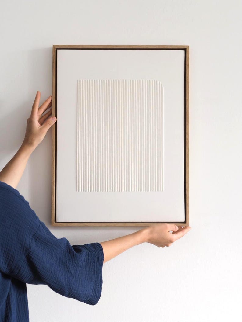 May include: A framed, abstract artwork with a white, textured surface featuring vertical and square patterns. The artwork is encased in a light brown wooden frame. The piece is held by a person wearing a dark blue top against a white wall.