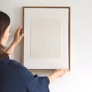 May include: A framed, textured white artwork with vertical lines and a rectangular shape. The artwork is set in a natural wood frame and is being held against a white wall. The piece has a minimalist, modern aesthetic.