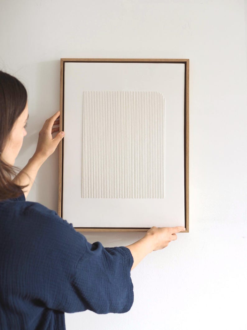 May include: A framed, textured white artwork with vertical lines and a rectangular shape. The artwork is set in a natural wood frame and is being held against a white wall. The piece has a minimalist, modern aesthetic.