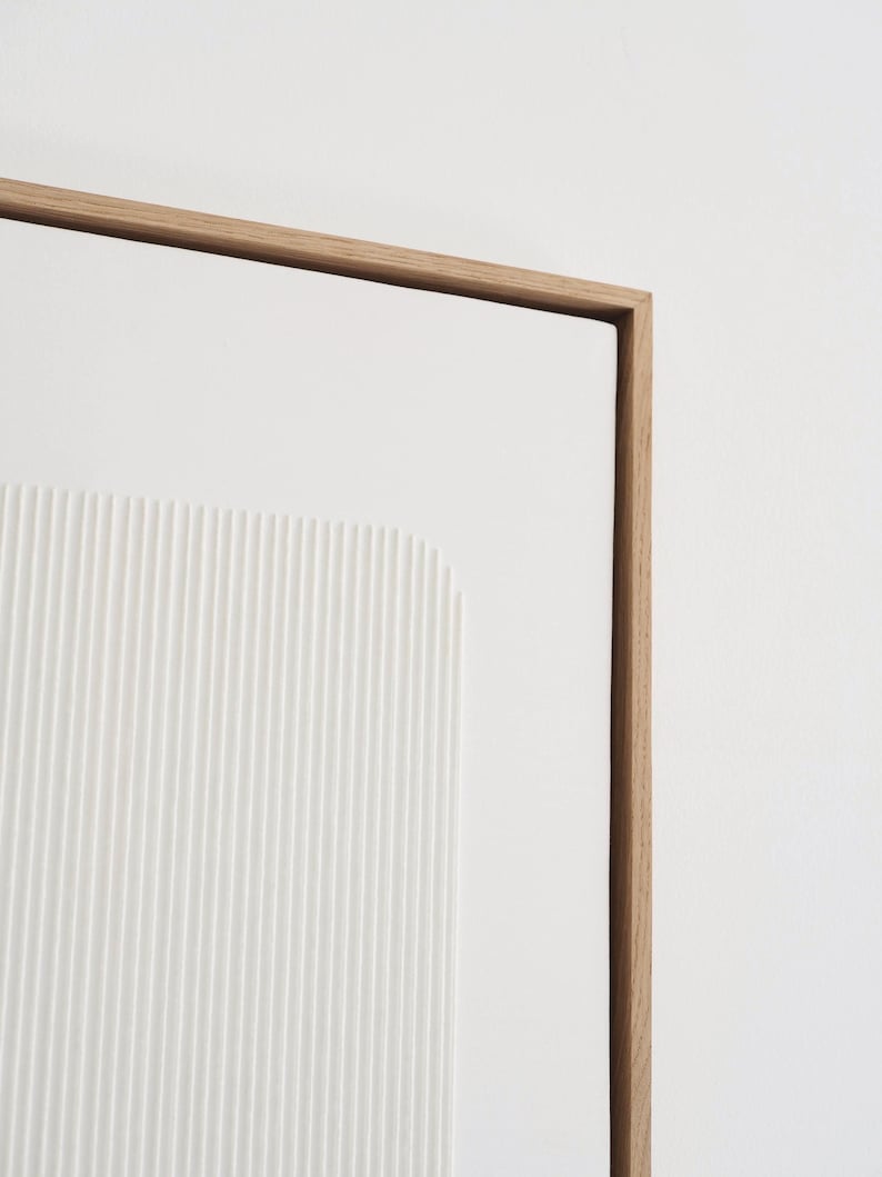May include: A modern, minimalist artwork featuring a white, textured panel with vertical lines and a rectangular shape. The artwork is framed in natural wood, creating a clean and contemporary aesthetic.