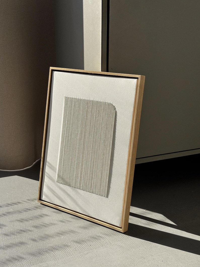 May include: A framed abstract art piece featuring a textured, off-white rectangular panel with vertical lines and a rounded top. The artwork is set within a light wood frame, creating a minimalist aesthetic.