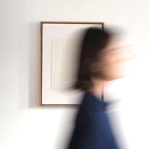 May include: A framed, minimalist artwork with a white, textured surface featuring vertical lines. The frame is a warm, light brown color. The artwork is mounted on a white wall. A person in a dark blue top is blurred in the foreground.