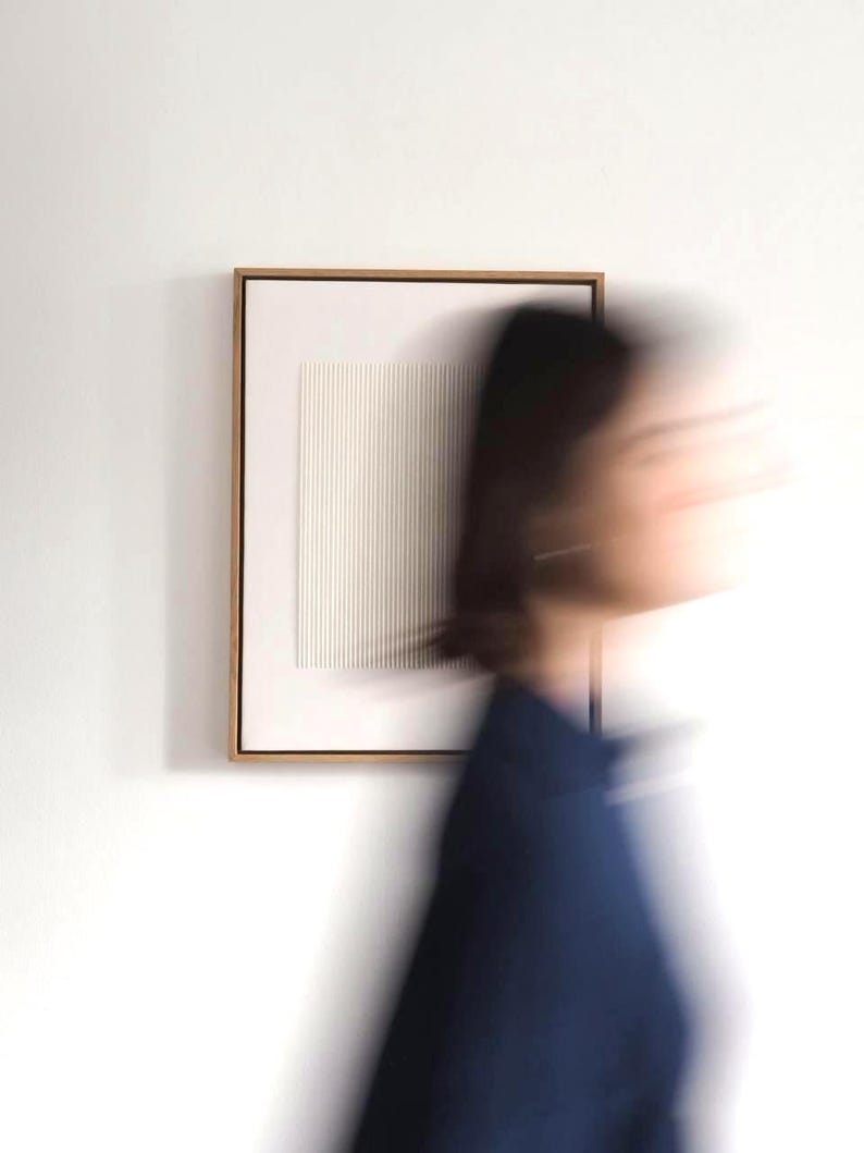May include: A framed, minimalist artwork with a white, textured surface featuring vertical lines. The frame is a warm, light brown color. The artwork is mounted on a white wall. A person in a dark blue top is blurred in the foreground.