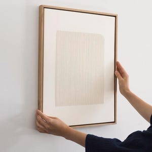 May include: A framed abstract art piece with a light wood frame. The artwork features a textured, off-white rectangular design with vertical lines. The piece is being held against a white wall.