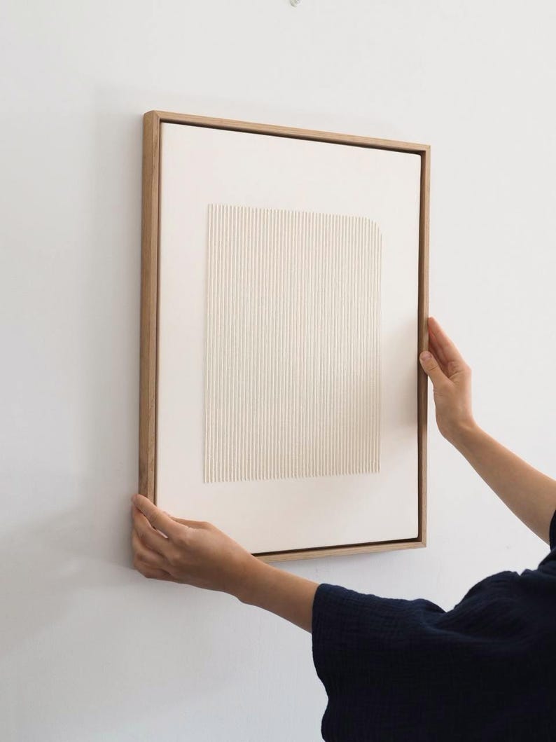 May include: A framed abstract art piece with a light wood frame. The artwork features a textured, off-white rectangular design with vertical lines. The piece is being held against a white wall.