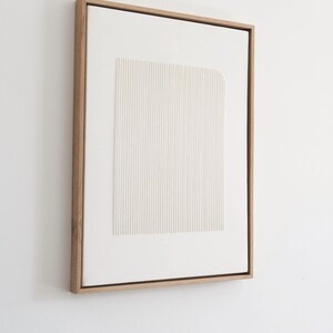 May include: A framed, abstract artwork featuring a white, textured rectangular design with vertical lines. The artwork is encased in a natural wood frame, creating a minimalist aesthetic. The piece is mounted on a white wall.
