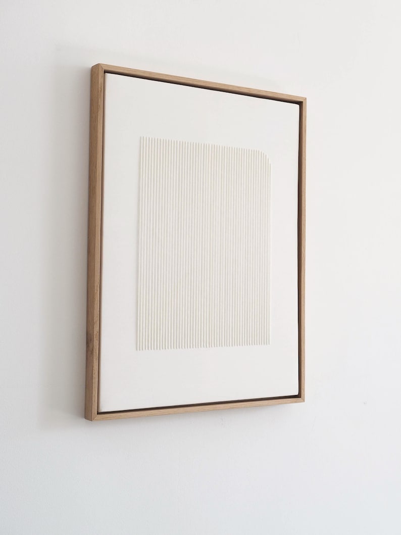 May include: A framed, abstract artwork featuring a white, textured rectangular design with vertical lines. The artwork is encased in a natural wood frame, creating a minimalist aesthetic. The piece is mounted on a white wall.