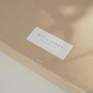 May include: Close-up of the back of a framed artwork. The artwork is mounted on a light brown cardboard backing. A white label with the text "STILL LINES MINIMALIST ART" is affixed to the cardboard. The frame is made of light-colored wood.