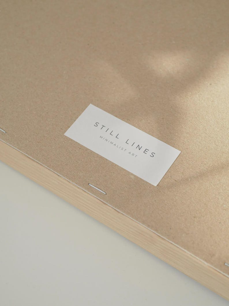 May include: Close-up of the back of a framed artwork. The artwork is mounted on a light brown cardboard backing. A white label with the text "STILL LINES MINIMALIST ART" is affixed to the cardboard. The frame is made of light-colored wood.