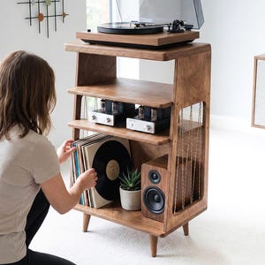 May include: A wooden record player stand with four tapered legs, featuring a top shelf for a turntable, a middle shelf holding two silver and black audio amplifiers, and a bottom shelf containing vinyl records, a small potted succulent, and a speaker.