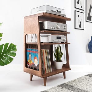 May include: A wooden media console with four tiers, featuring a top turntable, two silver audio components, and a bottom shelf for vinyl records. The side panels have vertical string detailing, and the unit stands on four tapered wooden legs.