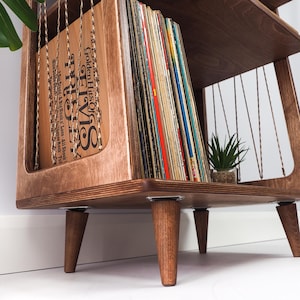 May include: A wooden record storage stand with four tapered legs, featuring a side panel with thin cord detailing and a collection of vinyl records. The front-most record sleeve is brown with black text reading Elvis Golden Hits.