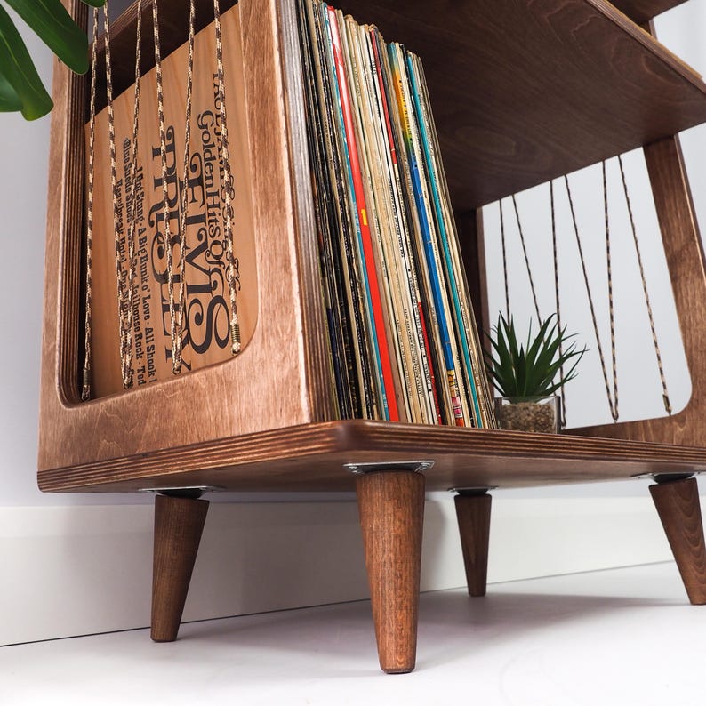May include: A wooden record storage stand with four tapered legs, featuring a side panel with thin cord detailing and a collection of vinyl records. The front-most record sleeve is brown with black text reading Elvis Golden Hits.