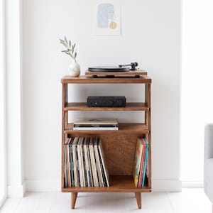 May include: A wooden record player stand with four tapered legs, featuring three open shelves. The top shelf holds a black turntable and a small vase with a green sprig. The middle shelf contains a black amplifier and two vinyl records.