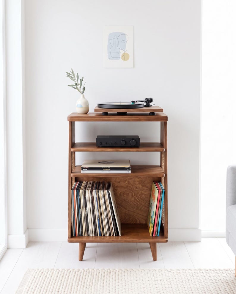 May include: A wooden record player stand with four tapered legs, featuring three open shelves. The top shelf holds a black turntable and a small vase with a green sprig. The middle shelf contains a black amplifier and two vinyl records.