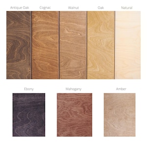 May include: A collection of eight wood finish samples arranged in a grid, labelled Antique Oak, Cognac, Walnut, Oak, Natural, Ebony, Mahogany, and Amber. Each rectangular sample displays a distinct wood grain pattern and colour tone ranging from light beige to dark brown and deep purple-black.