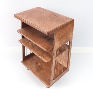 May include: A brown wooden shelving unit featuring a flat top surface and two lower shelves. The side panels are constructed with a cutout design, accented by thin, vertical, light-coloured cord detailing that connects the top to the base.
