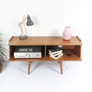 Peut inclure: Meuble TV en bois de style milieu du si&egrave;cle avec une finition marron clair. Il comprend une &eacute;tag&egrave;re ouverte avec un r&eacute;cepteur st&eacute;r&eacute;o argent&eacute;, des livres et un vase rose. Une lampe de bureau grise avec un bras en bois est pos&eacute;e dessus.