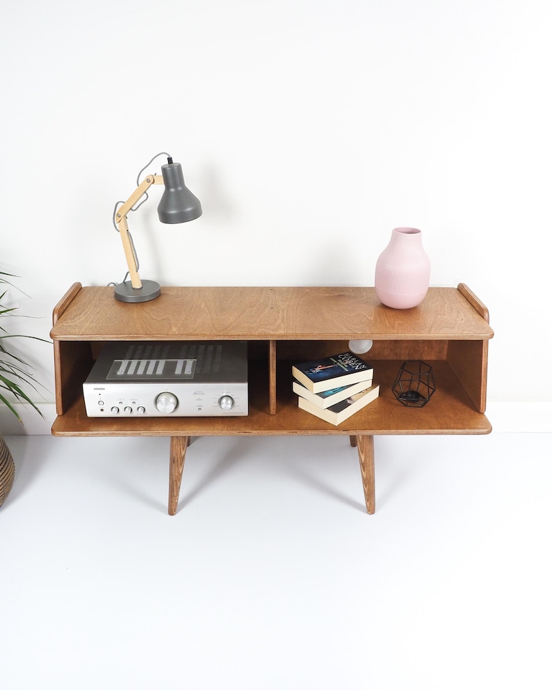 Peut inclure: Meuble TV en bois de style milieu du si&egrave;cle avec une finition marron clair. Il comprend une &eacute;tag&egrave;re ouverte avec un r&eacute;cepteur st&eacute;r&eacute;o argent&eacute;, des livres et un vase rose. Une lampe de bureau grise avec un bras en bois est pos&eacute;e dessus.