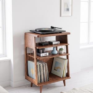 May include: A dark wood record player stand with four tapered legs, featuring a top surface for a turntable, two middle shelves for audio equipment and decor, and a bottom section for vinyl record storage with vertical side support wires.
