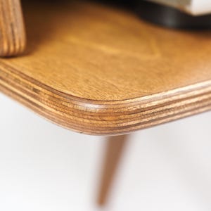 Peut inclure: Gros plan sur un coin de table en bois. La table a une finition brun clair avec un grain de bois visible et un bord arrondi. La construction en couches de la table est &eacute;vidente, et un pied est visible.