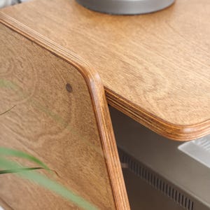 Peut inclure: Gros plan sur une table d'appoint en bois avec un fini grain de bois naturel. La table a des bords arrondis et une construction en couches. Une base de lampe grise est pos&eacute;e sur le dessus. Le design de la table est moderne et minimaliste.