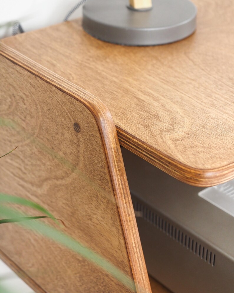 Peut inclure: Gros plan sur une table d'appoint en bois avec un fini grain de bois naturel. La table a des bords arrondis et une construction en couches. Une base de lampe grise est pos&eacute;e sur le dessus. Le design de la table est moderne et minimaliste.