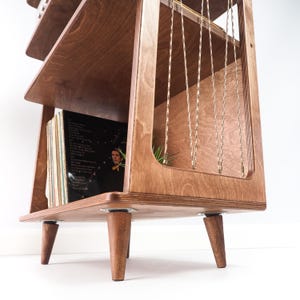 May include: A wooden record storage unit with a tiered design and tapered legs. The unit features multiple shelves for vinyl records, with a side panel containing vertical gold-toned metal chains. The wood has a visible grain and warm brown finish.
