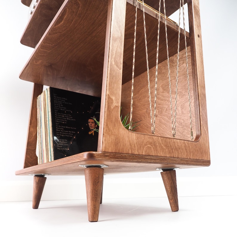 May include: A wooden record storage unit with a tiered design and tapered legs. The unit features multiple shelves for vinyl records, with a side panel containing vertical gold-toned metal chains. The wood has a visible grain and warm brown finish.