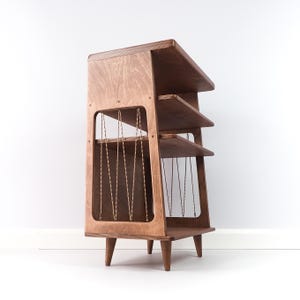 May include: A wooden shelving unit with a light brown finish, featuring three horizontal tiers and four tapered legs. The side panels have large rectangular cutouts with a zigzag pattern of thin, twisted cord running vertically between the shelves.