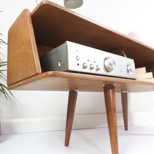 Peut inclure: Meuble st&eacute;r&eacute;o en bois de style moderne du milieu du si&egrave;cle, avec un r&eacute;cepteur argent&eacute;. Le meuble a un dessus rectangulaire, des c&ocirc;t&eacute;s inclin&eacute;s et des pieds effil&eacute;s. Une petite lampe est pos&eacute;e au-dessus.