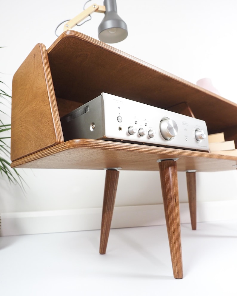 Peut inclure: Meuble st&eacute;r&eacute;o en bois de style moderne du milieu du si&egrave;cle, avec un r&eacute;cepteur argent&eacute;. Le meuble a un dessus rectangulaire, des c&ocirc;t&eacute;s inclin&eacute;s et des pieds effil&eacute;s. Une petite lampe est pos&eacute;e au-dessus.