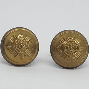 Puede incluir: Dos botones antiguos de latón con un emblema detallado que presenta una corona, una cruz y patrones intrincados. Los botones son redondos y tienen una apariencia ligeramente envejecida, lo que sugiere que son vintage.